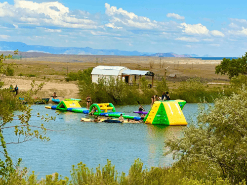 People enjoying inflatable water activities on a lake, with a backdrop of mountains and a clear blue sky.